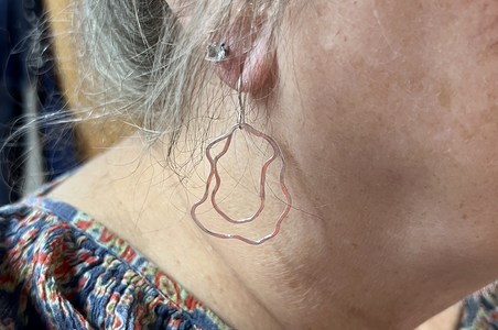 A close-up of a student's ear wearing a handmade wavy wire hoop earring in a sterling silver, with gray hair and a patterned top visible.