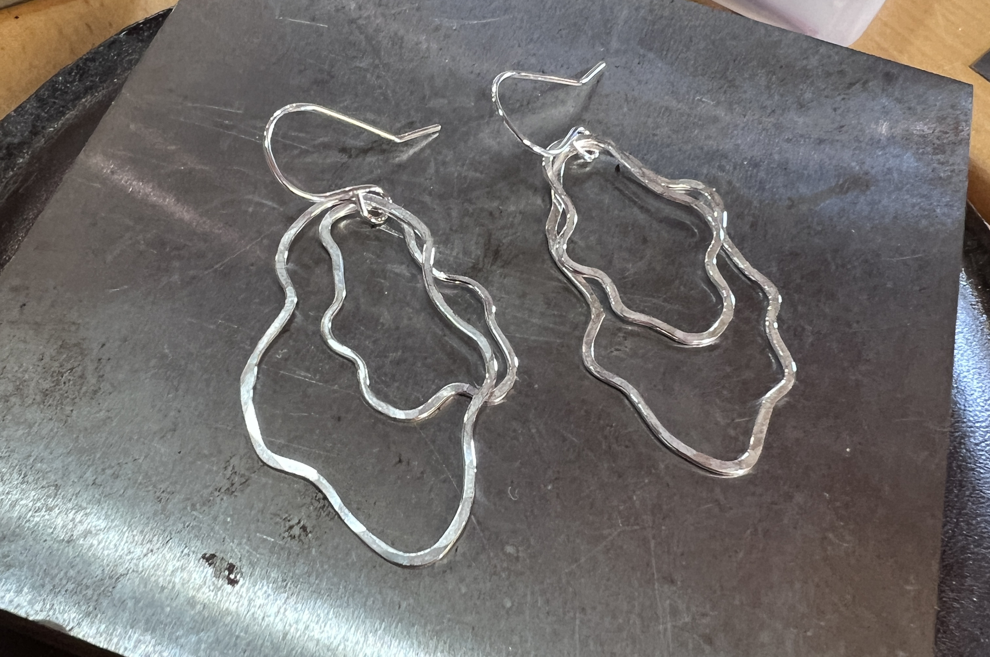 A pair of sterling silver wavy abstract hoop earrings with fishhook earwires, displayed on a steel bench block