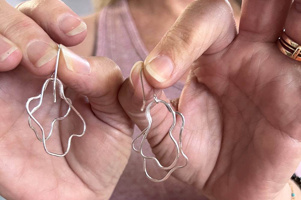 A close-up of a person holding a pair of silver wavy abstract hoop earrings with fishhook backs between their hands.