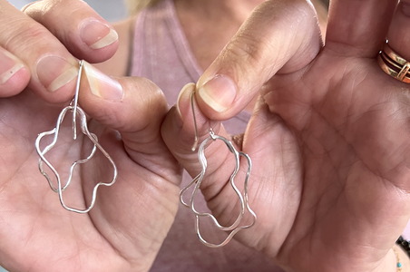 A close-up of a person holding a pair of silver wavy abstract hoop earrings with fishhook backs between their hands.