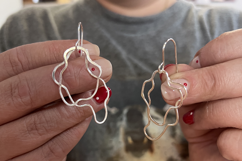 A student holding a pair of abstract wavy wire hoop earrings with fishhook backings. 