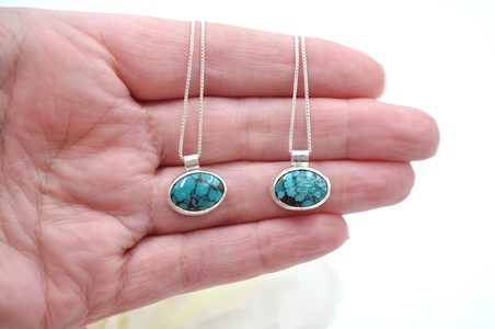 Two turquoise stone pendants on silver chains held in a hand against a white background