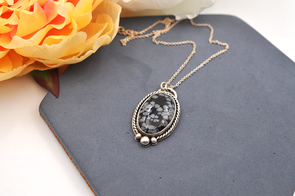 Necklace with a black and gray snowflake obsidian pendant on a gray surface next to yellow flowers