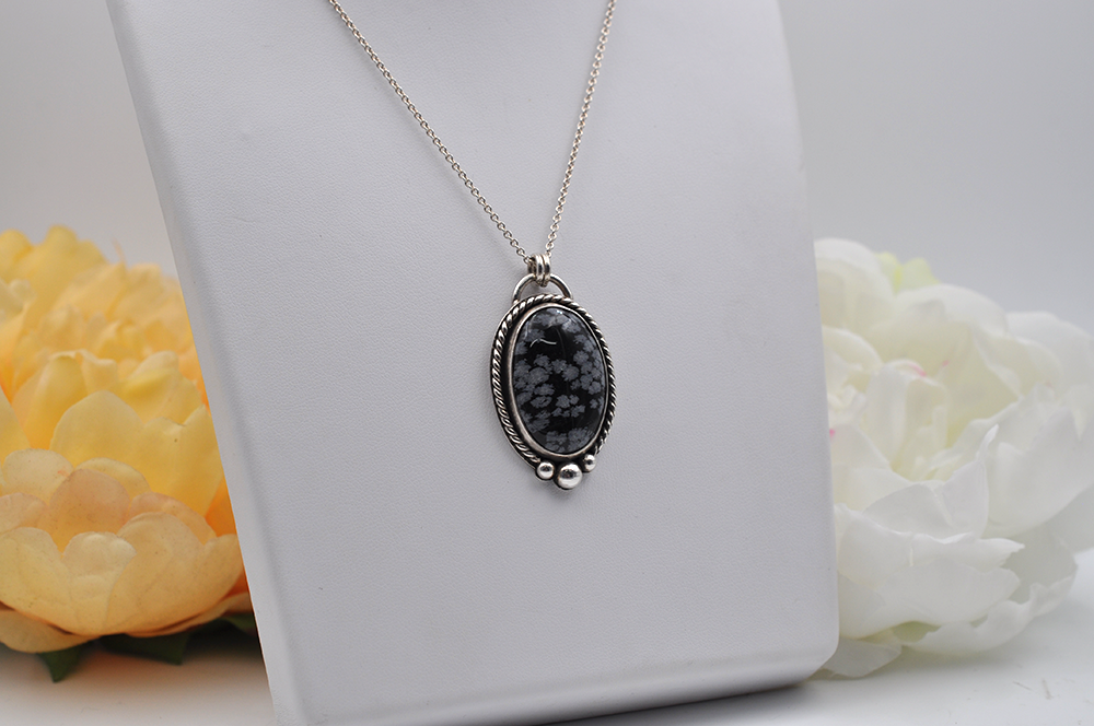 Necklace with a black and gray stone pendant set in sterling silver on a white stand with flowers in the background