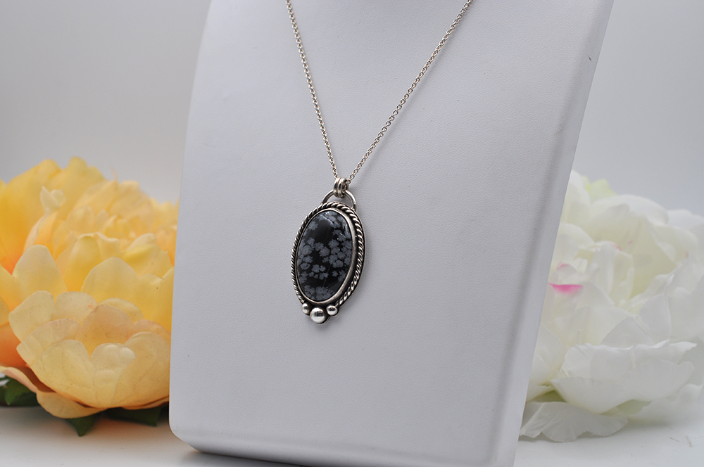 Black and gray floral snowflake obsidian  pendant necklace set in sterling silver on a white stand with flowers in the background