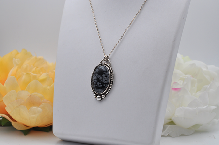 Black and gray floral snowflake obsidian  pendant necklace set in sterling silver on a white stand with flowers in the background