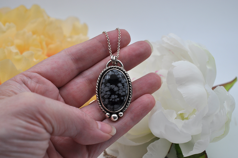 Hand holding a necklace with a black and gray snowflake obsidian pendant set in sterling silver against a floral background