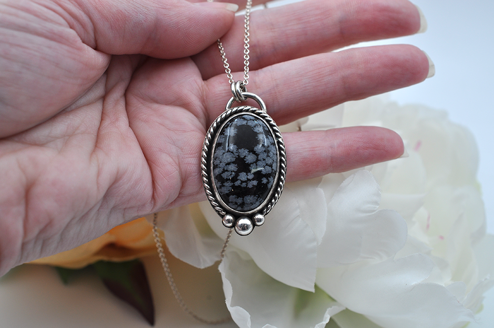 Silver necklace with an black and gray snowflake obsidian oval pendant held by a hand against a white flower background