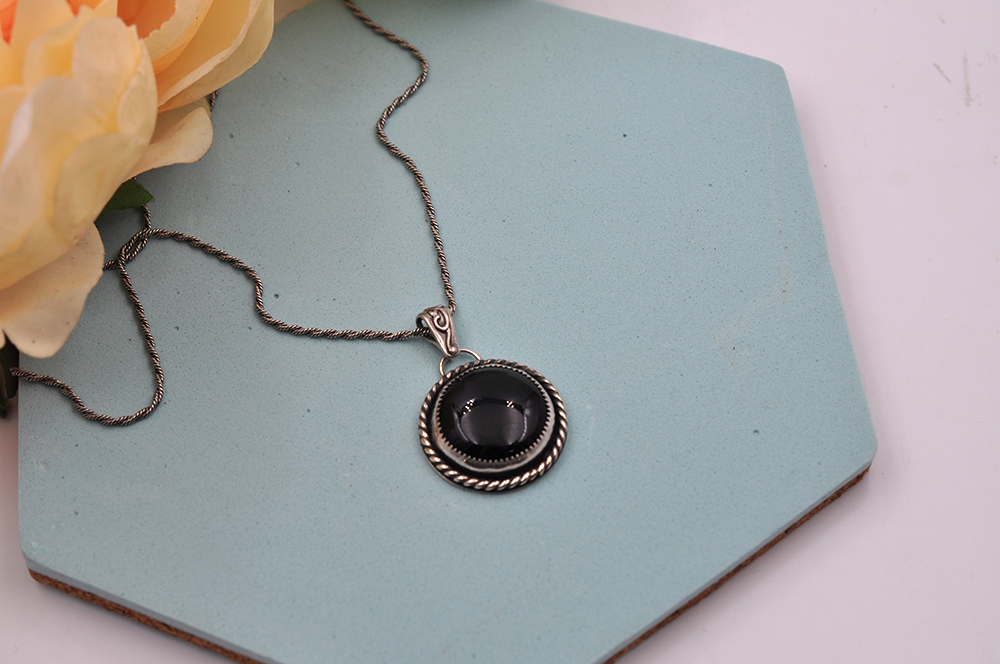 Necklace with a black onyx pendant on a light blue hexagonal stand with flowers in the background