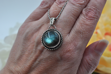 Hand wearing a silver necklace with a greenish-blue pendant against a blurred background