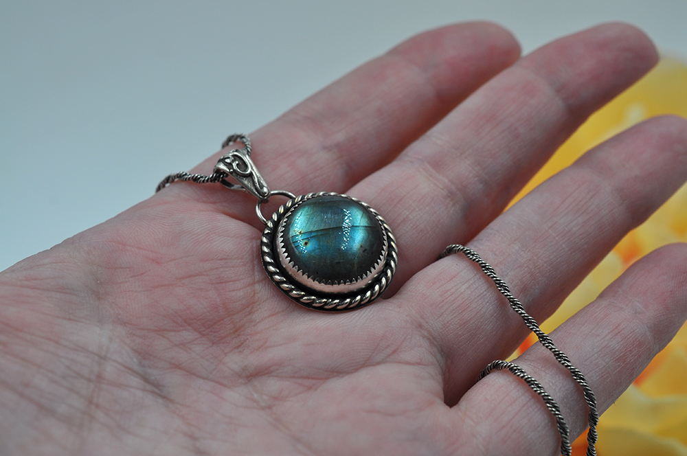 Hand holding a silver pendant with a greenish-blue labradorite gemstone against a blurred background