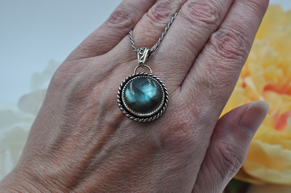 Hand holding a silver pendant with a green labradorite gemstone against a blurred floral background