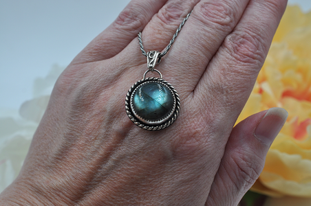 Hand holding a silver pendant with a green labradorite gemstone against a blurred floral background
