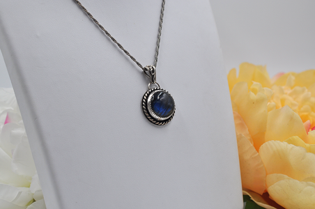 Silver necklace with a blue labradorite stone pendant on a white surface in front of yellow flowers.