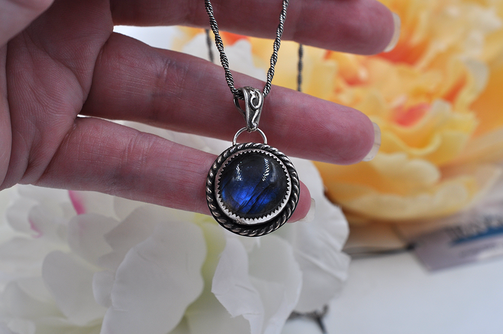 Hand holding a necklace with a blue labradorite pendant against a blurred floral background