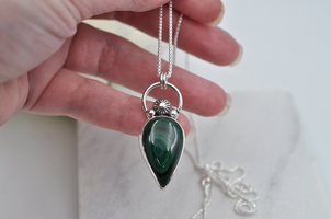 Hand holding a silver necklace with a green teardrop pendant against a white background