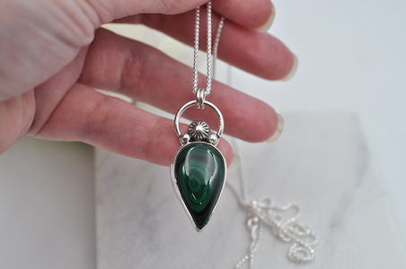 Hand holding a silver necklace with a green teardrop pendant against a white background