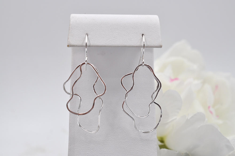 Silver abstract earrings on a white stand with white flowers in the background