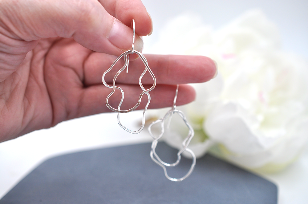 Hand holding an organic silver wire earrings with a blurred white flower in the background
