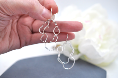 Hand holding an organic silver wire earrings with a blurred white flower in the background