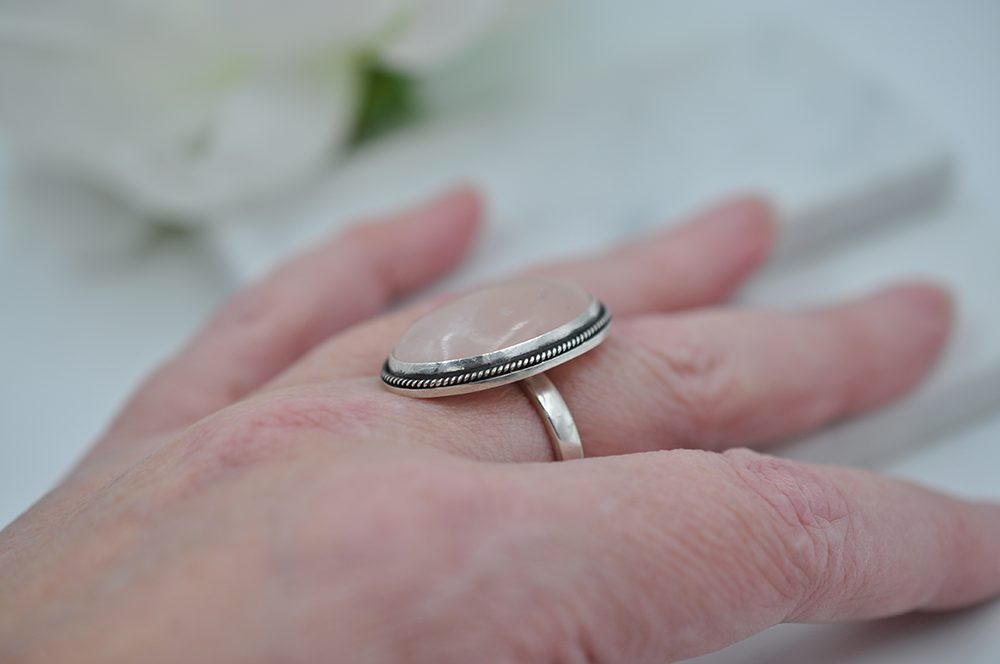 Silver ring on a person's finger showing the twisted rope detail around the stone. The ring is pictured in front of a blurred floral background
