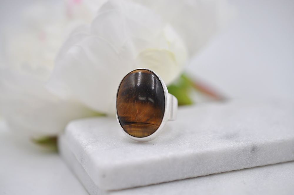 Silver ring with a large tiger's eye brown stone on a marble surface with flowers in the background