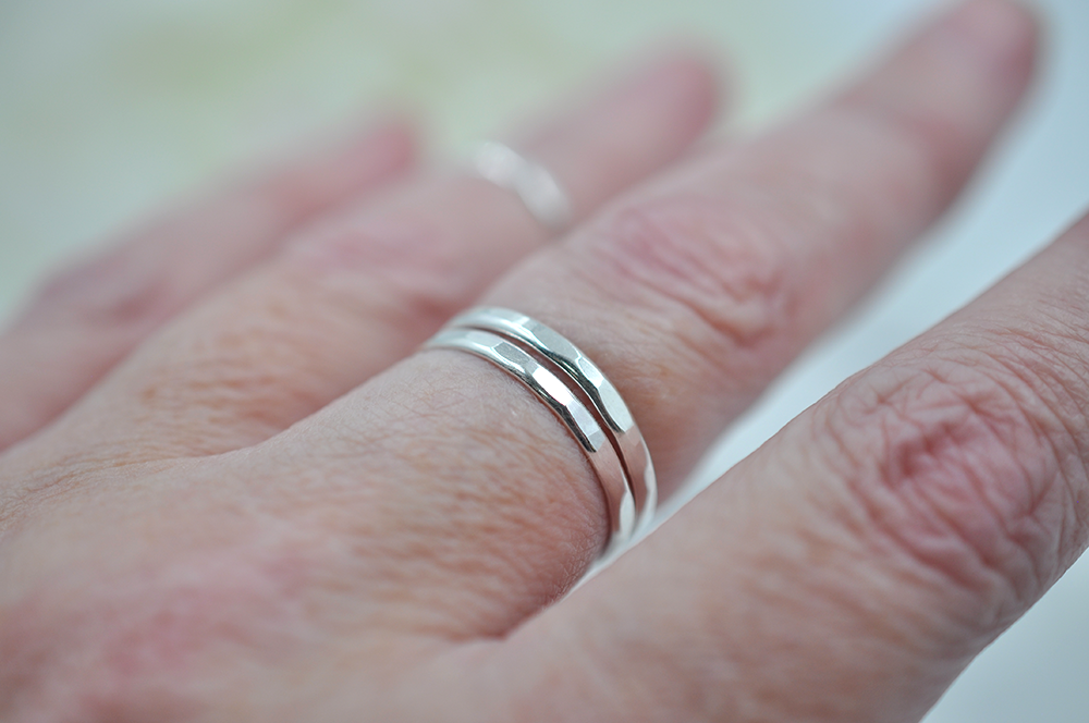 Close-up of a hand wearing two hammered silver rings stacked together on the middle finger pictured in front of a blurred background