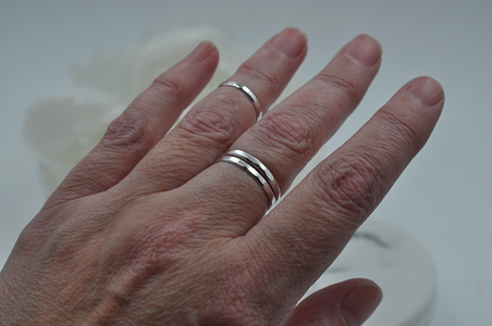 Hand wearing two silver hammered rings stacked on middle finger and one smaller ring on the ring finger above the first knuckle pictured in front of a neutral background