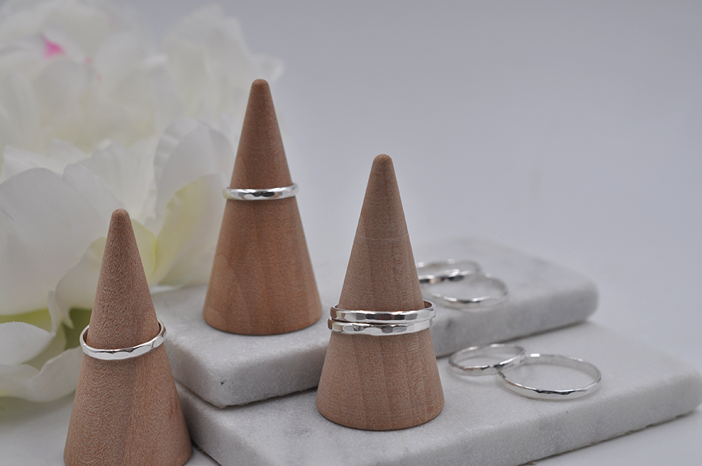 Three wooden cone-shaped ring stands with hammered silver rings on a marble surface with flowers in the background.