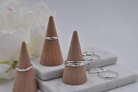 Three wooden cone-shaped ring stands with hammered silver rings on a marble surface with flowers in the background.