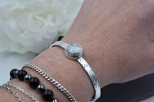 Silver cuff bracelet with a hammered texture and gray and white round stone shown on a wrist with a white flower in the background