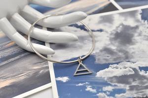 Pictured here resting on the fingers of a white mannequin hand for scale is a round sterling silver bangle bracelet with a small air charm hanging from the continuous circle. Air symbol in a triangle with a line through the center. 