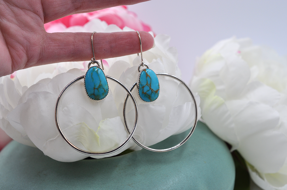 A pair of sterling silver hoop earrings with natural turquoise mountain turquoise stones, displayed against a floral background.