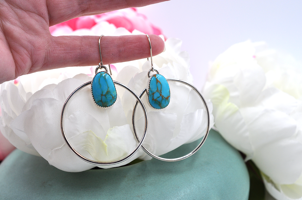 A pair of sterling silver hoop earrings with natural turquoise mountain turquoise stones, displayed against a floral background.