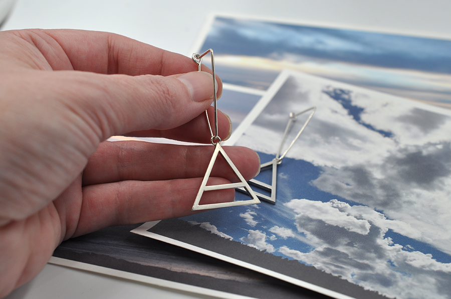Featured here is a pair of sterling silver triangular drop hoops with a large air charm hanging from the bottom point of the triangle. The air symbol featured here is a triangle with a line through the center. 