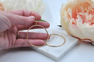 Large gold drop hoops pictured in hand for scale with another hoop earring pictured laying flat on a marble slab. 