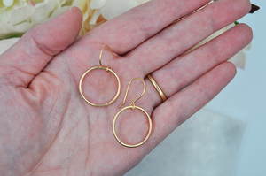 A pair of small gold drop hoops pictured in the palm of a models hand for scale.