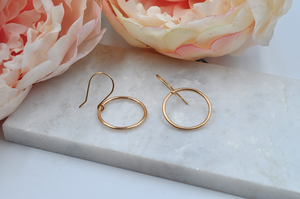 Gold round drop hoop earrings that measure approximately one inch in width pictured laying on a marble slab. 