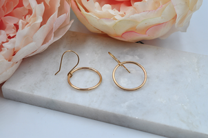 Gold round drop hoop earrings that measure approximately one inch in width pictured laying on a marble slab. 