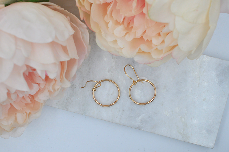 Gold round drop hoop earrings that measure approximately one inch in width pictured laying on a marble slab. 